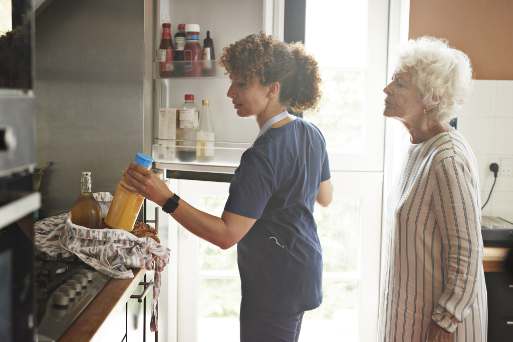 Mature female nurse keeping bottle in refrigerator standing by senior woman in kitchen at home